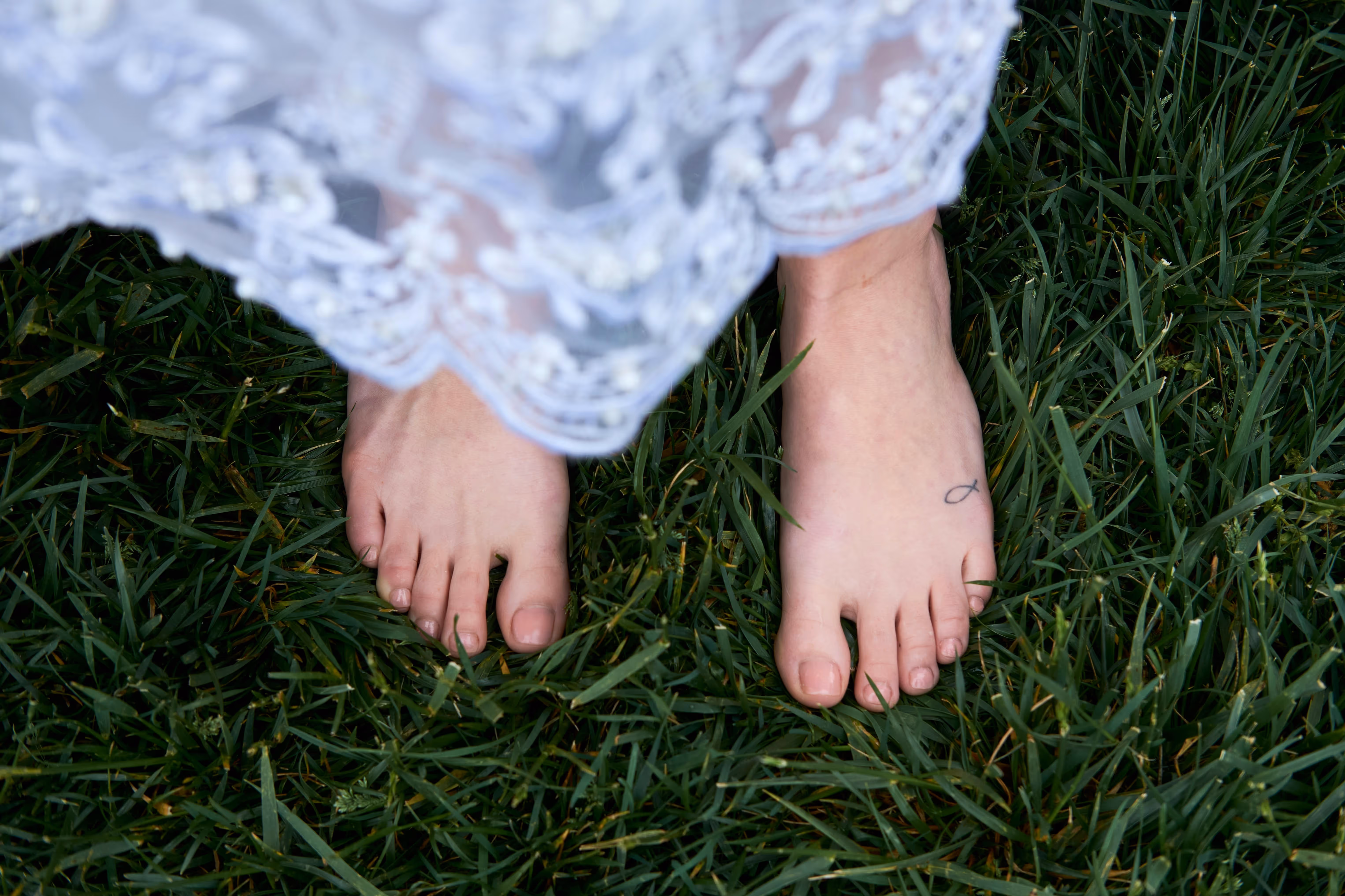 A pair of bare feet in grass with a lace white dress in blowing in the wind.
