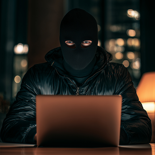 Person wearing a black ski mask and jacket using a laptop in a dimly lit room at night.