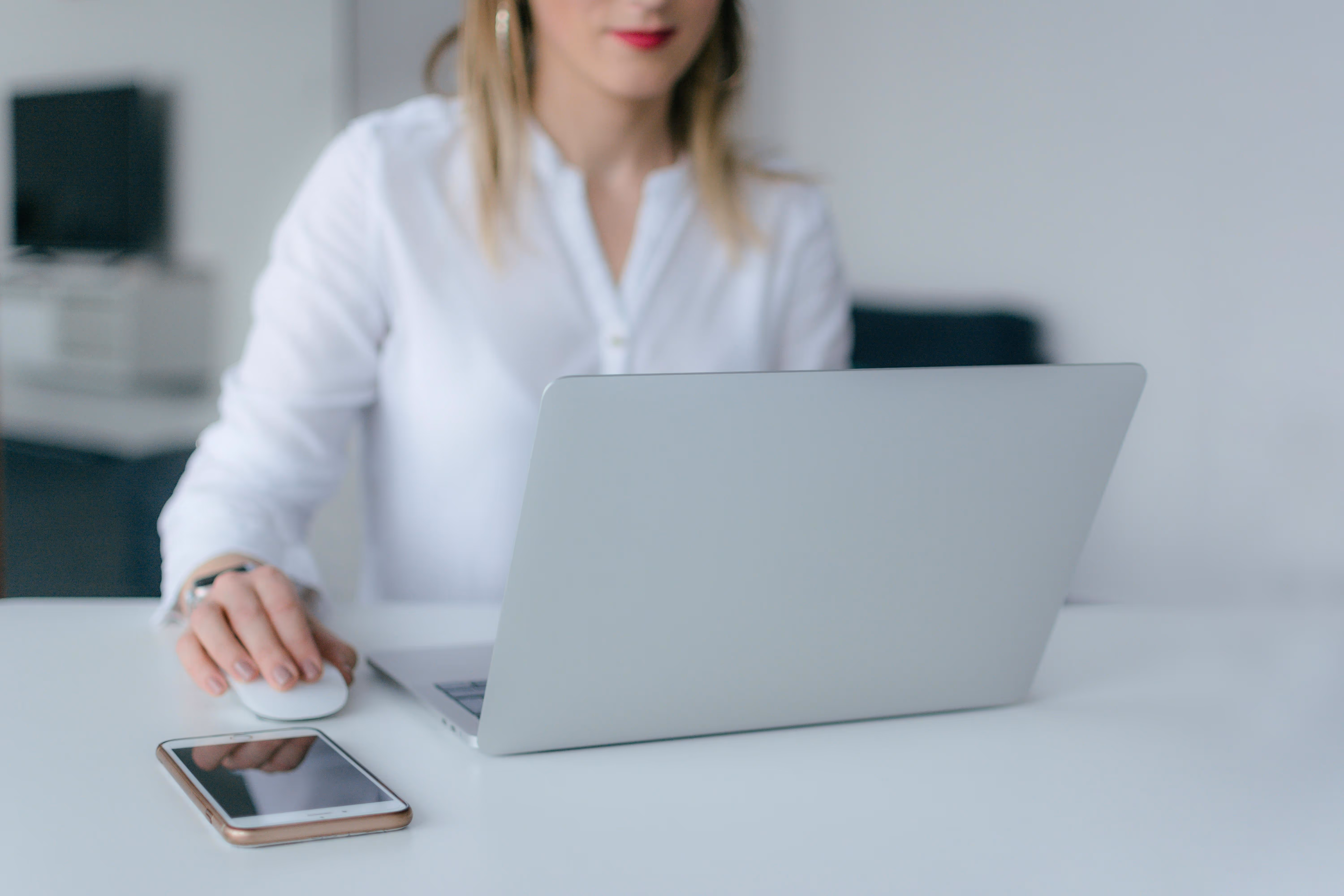 Woman in a white blouse using a silver laptop with a smartphone placed on the desk.