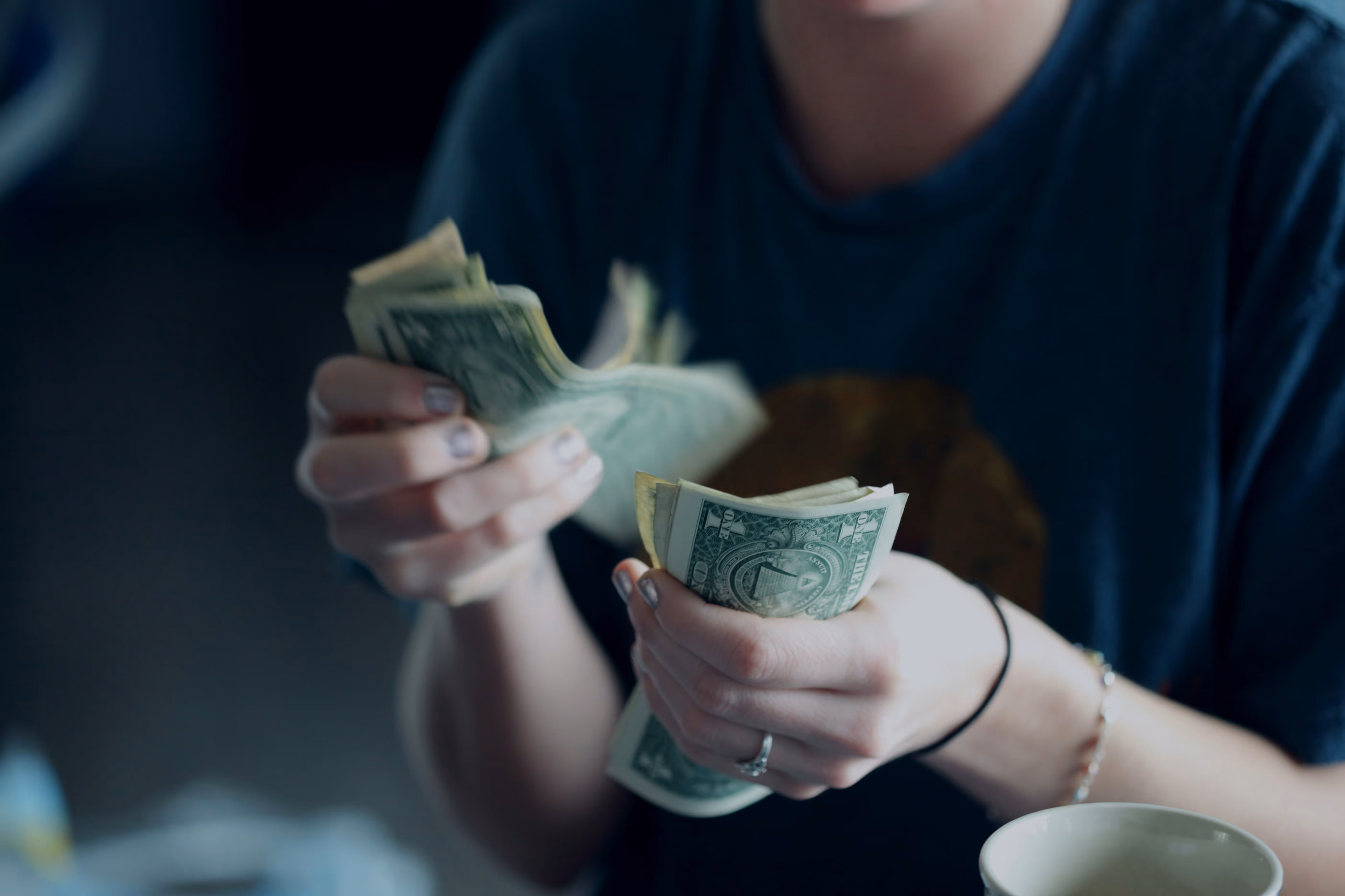 Person counting US one-dollar bills with both hands, wearing a dark shirt and bracelet.