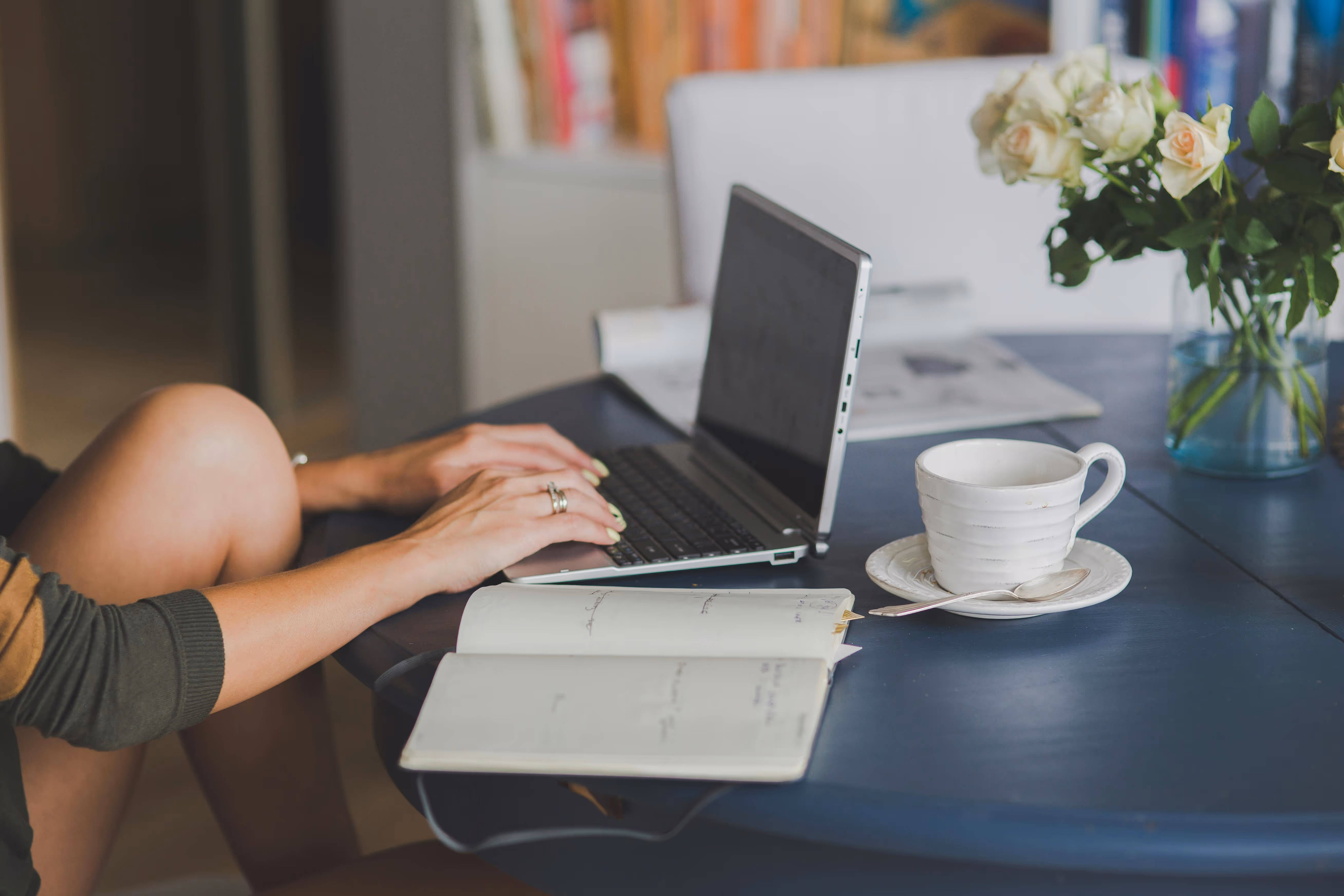 Person typing on a laptop at a blue table with an open notebook, white coffee cup on a saucer with spoon, and a vase of white roses.