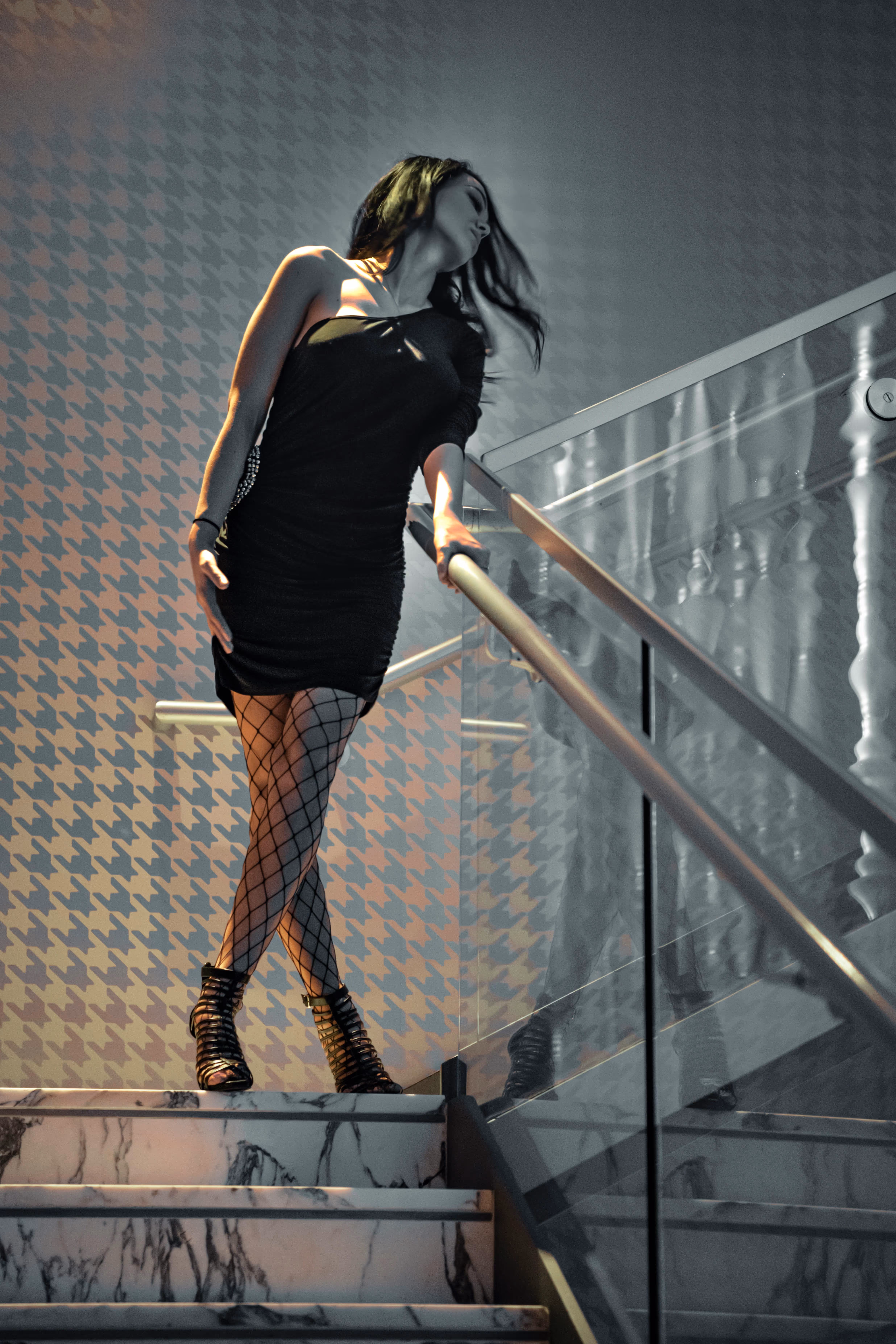 Woman in black dress and fishnet stockings standing on marble stairs, holding glass railing with patterned wall behind.