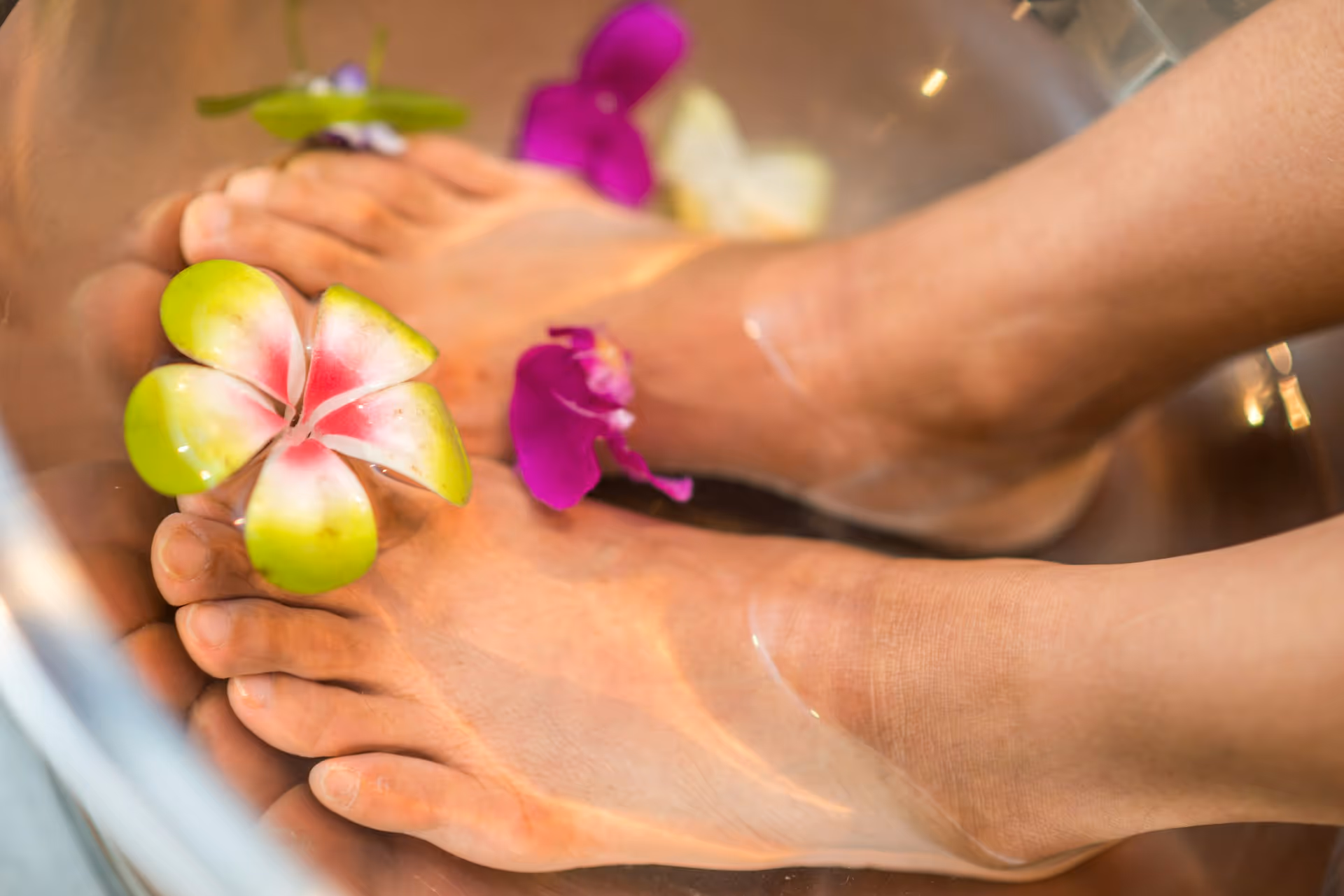 Feet soaking in water with green and pink frangipani and purple orchid flowers floating around.