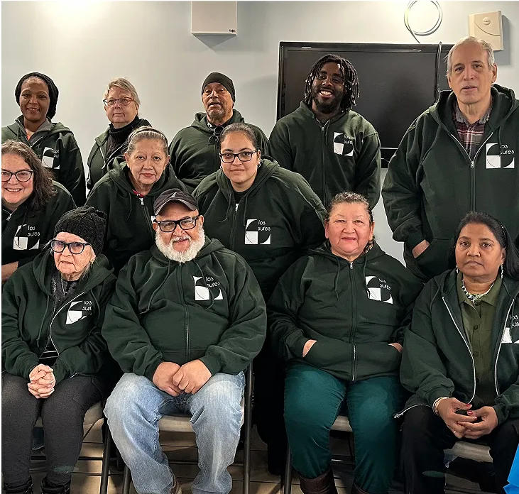 A diverse group of people wearing matching dark green hoodies with a white 'los sures' logo.