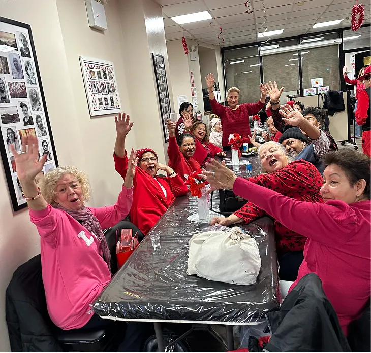 Group of older adults happily waving and sitting around a long table in a decorated room.