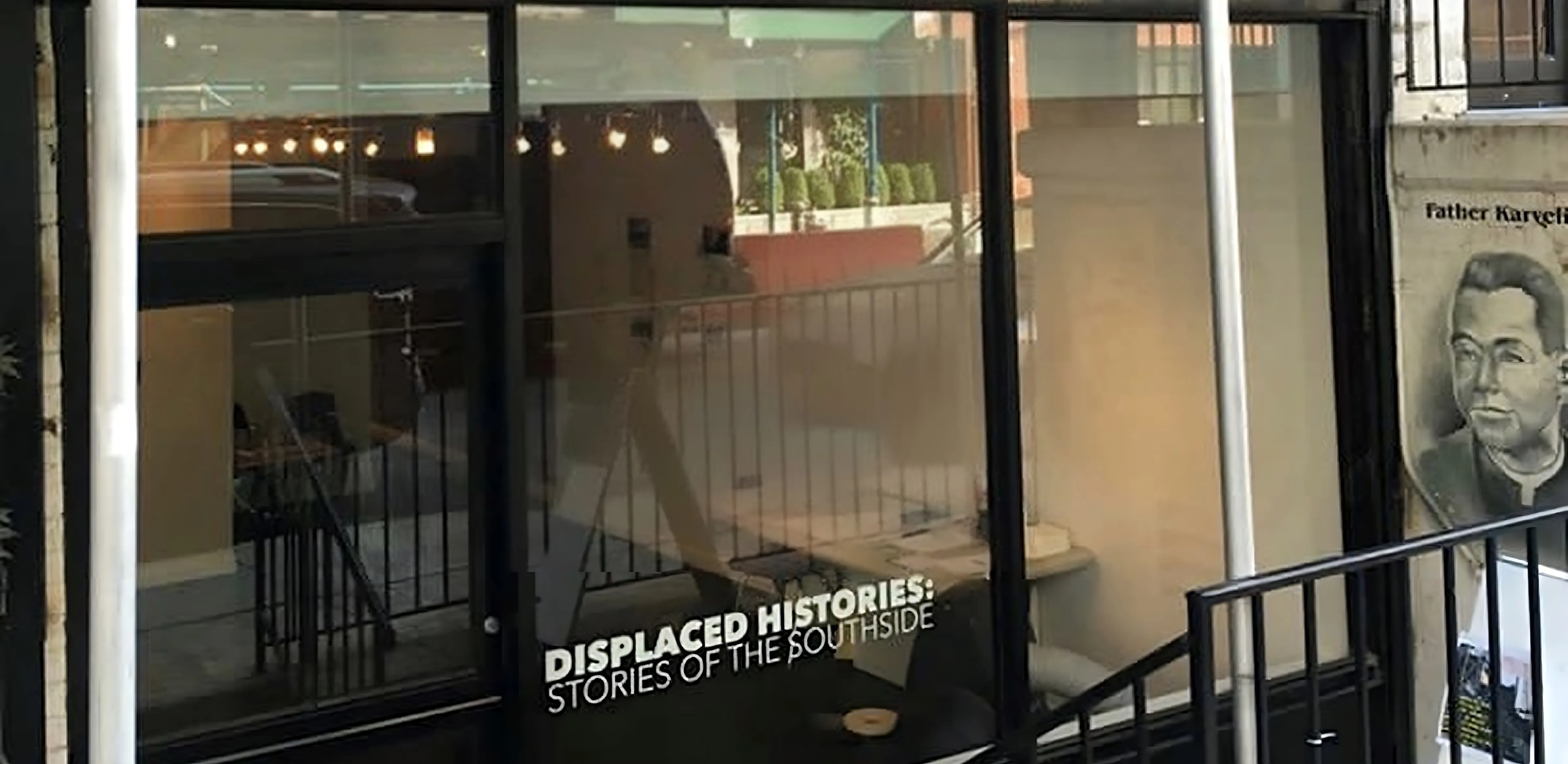 Storefront window displaying "Displaced Histories: Stories of the Southside" exhibit title, with reflections of the street and a portrait mural of Father Kargel visible on the building's exterior.