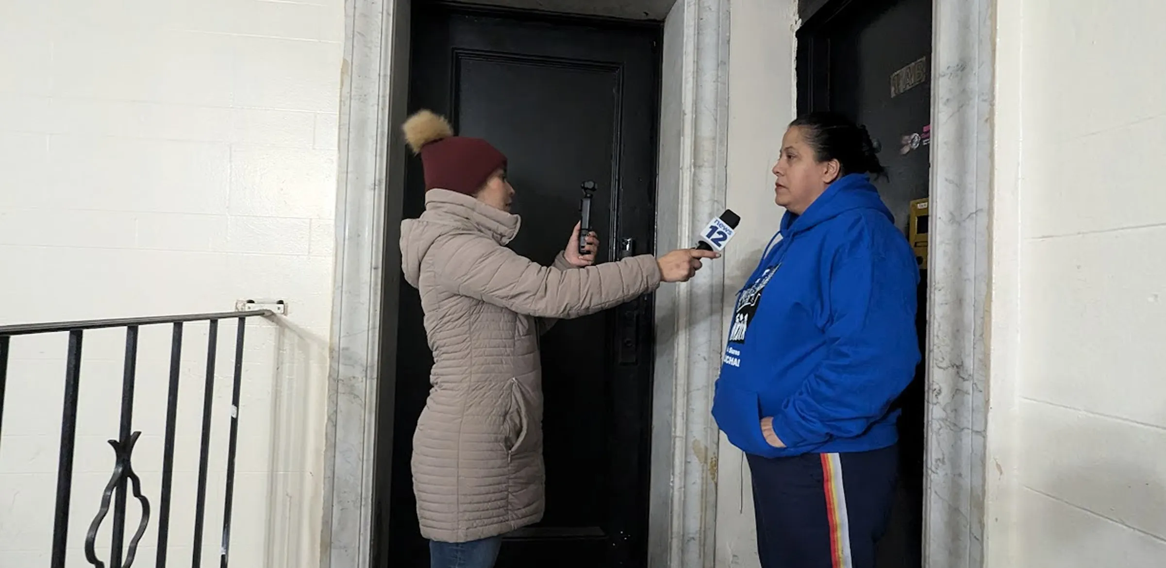Reporter in a beige coat holding a microphone interviewing a woman in a blue hoodie.