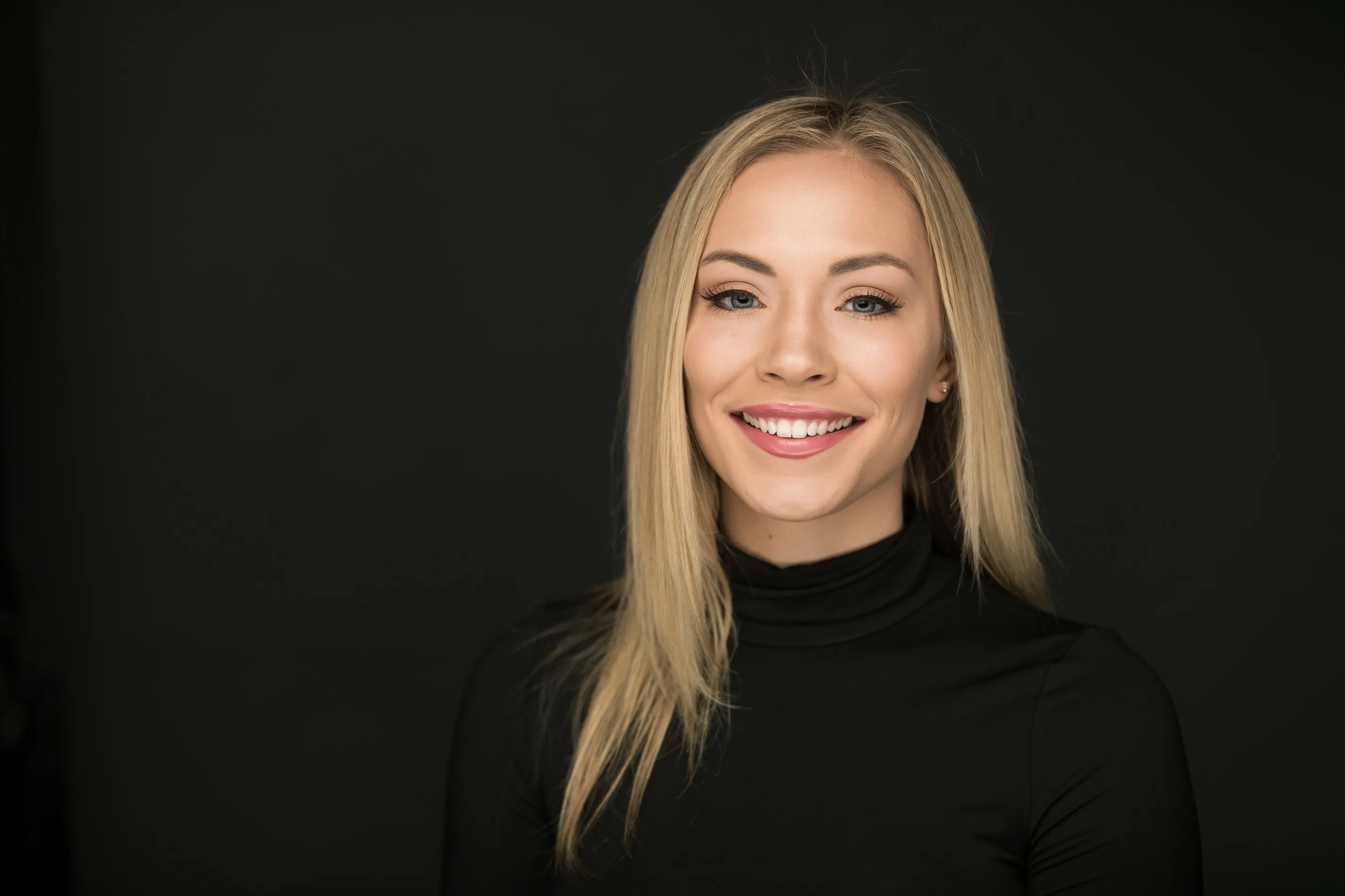 Smiling blonde woman with straight hair wearing a black turtleneck against a dark background at Legacy Dental Pasadena after getting her Invisalign.