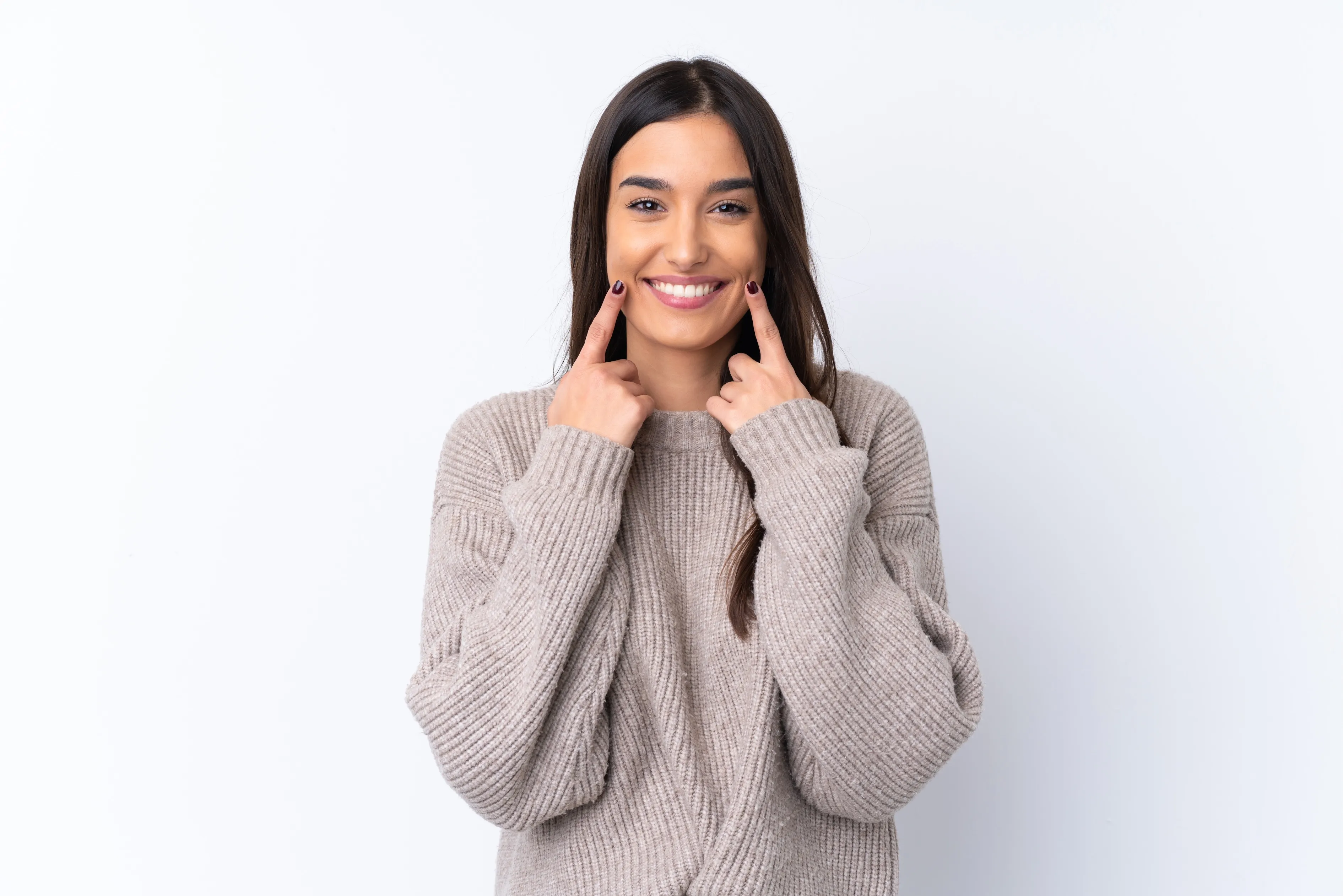 Smiling woman in a beige sweater pointing to her smile with both index fingers after getting ICON Resin Infiltration at Legacy Dental Pasadena