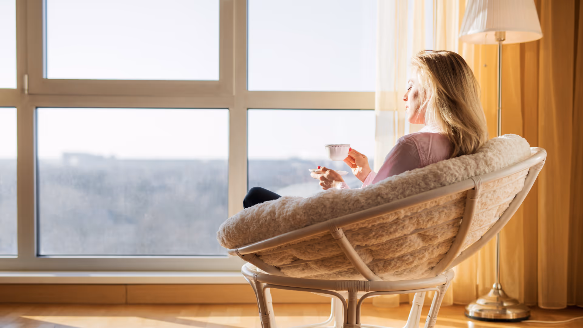 Woman sitting in a cozy chair by a large window, holding a cup and saucer, bathed in warm sunlight.