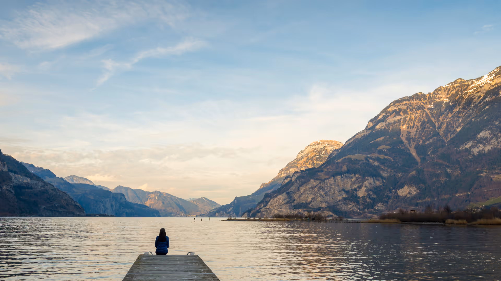 Person sitting alone at the end of a wooden dock overlooking a large lake surrounded by mountains under a partly cloudy sky.