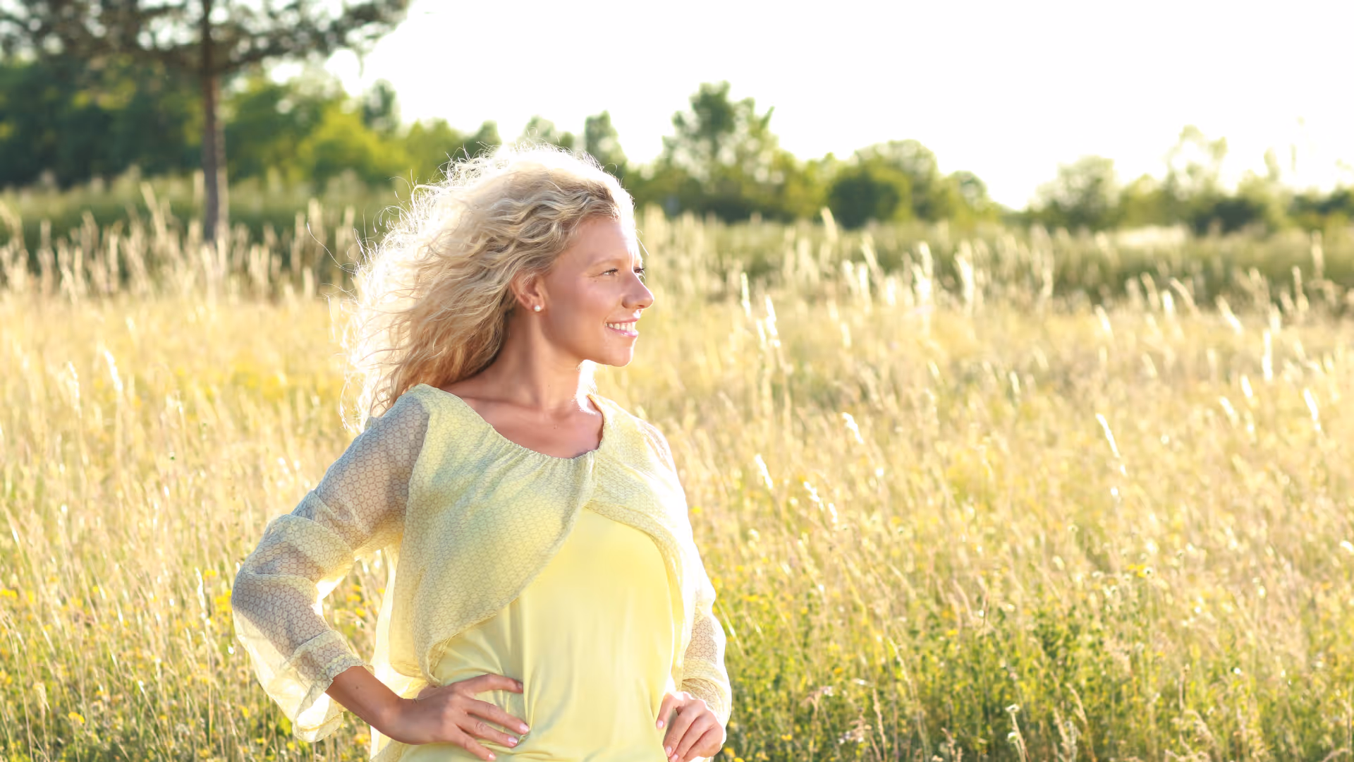 Smiling woman with curly blonde hair standing in a sunlit grassy field, wearing a sheer yellow top.