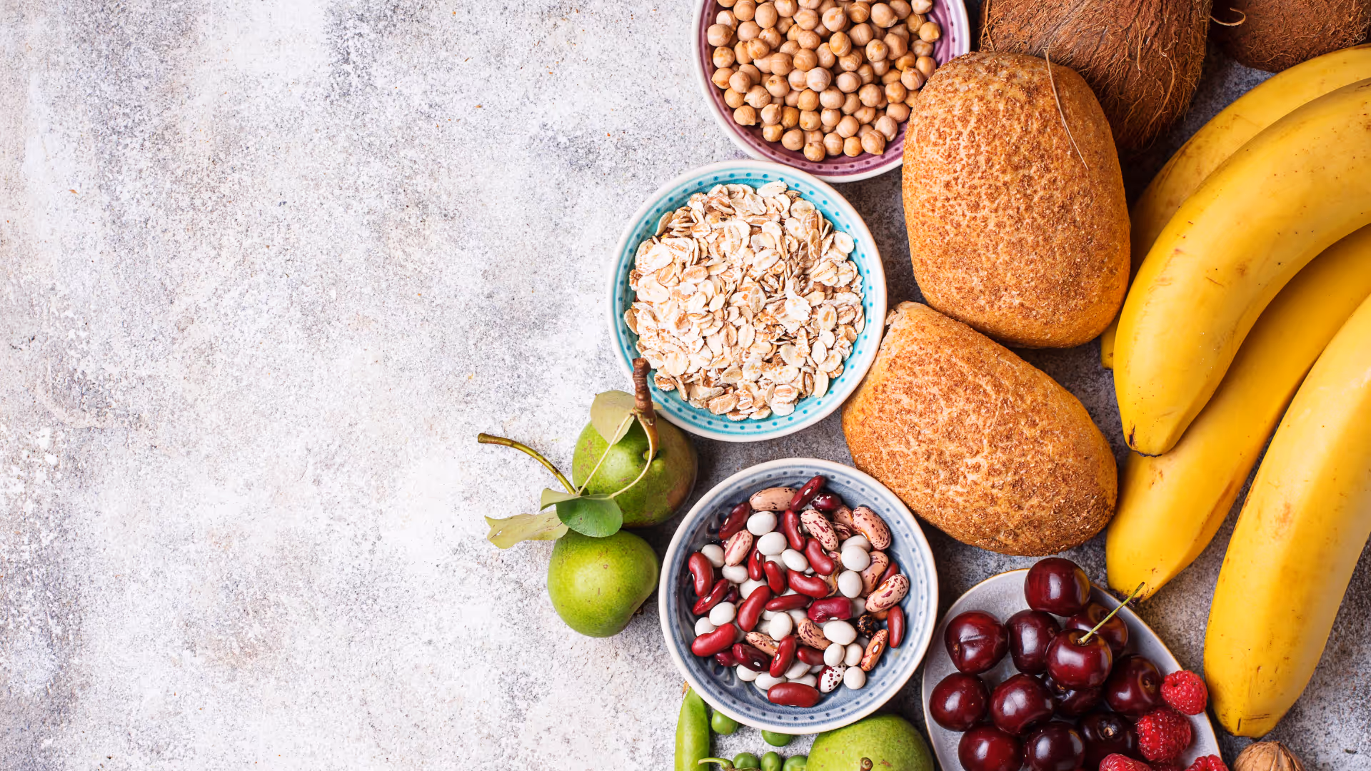 Assortment of healthy foods including bowls of oats, beans, chickpeas, whole grain bread, bananas, cherries, raspberries, green pears, peas, and whole coconuts.