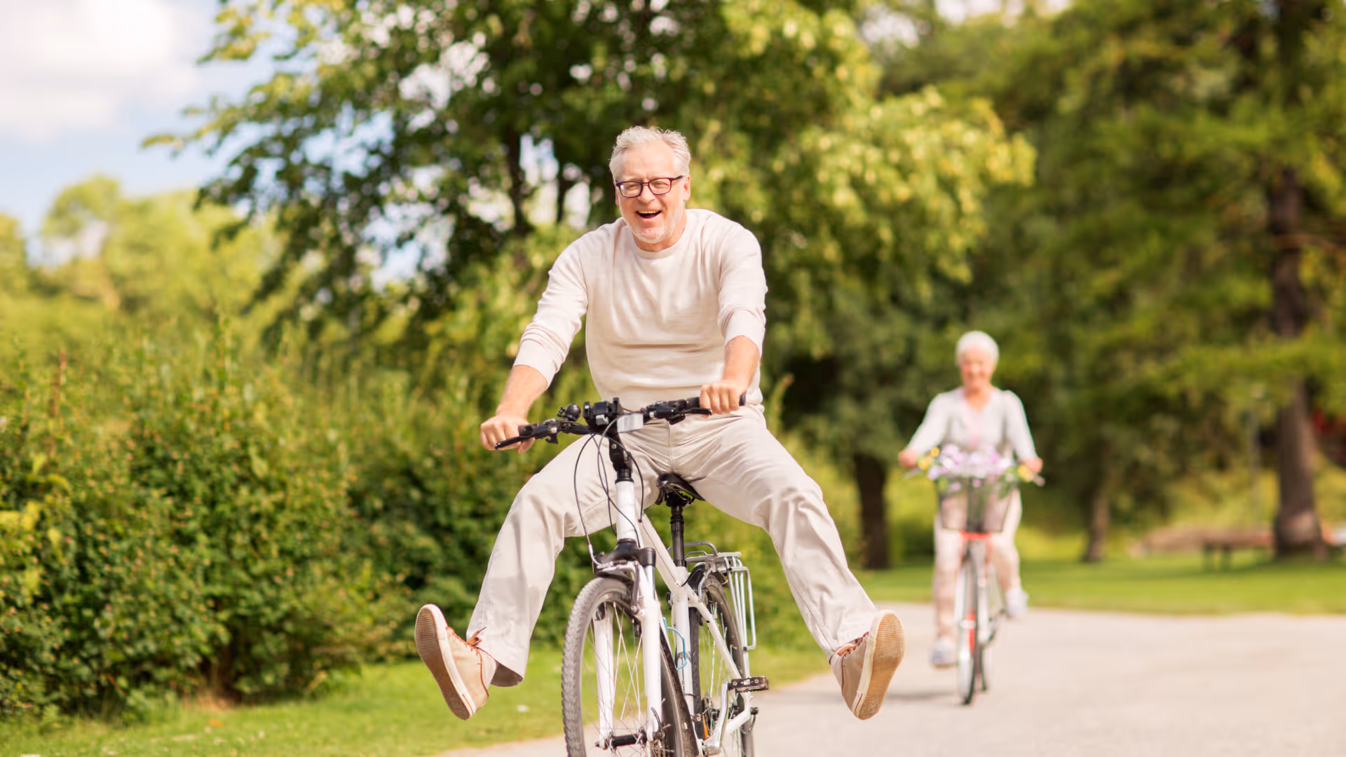 Smiling elderly man riding a bike with legs extended forward on a park path, with an elderly woman cycling behind him.