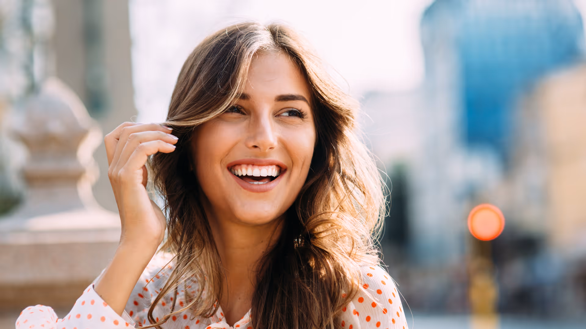 Smiling young woman with long wavy hair wearing a white shirt with orange polka dots outdoors.