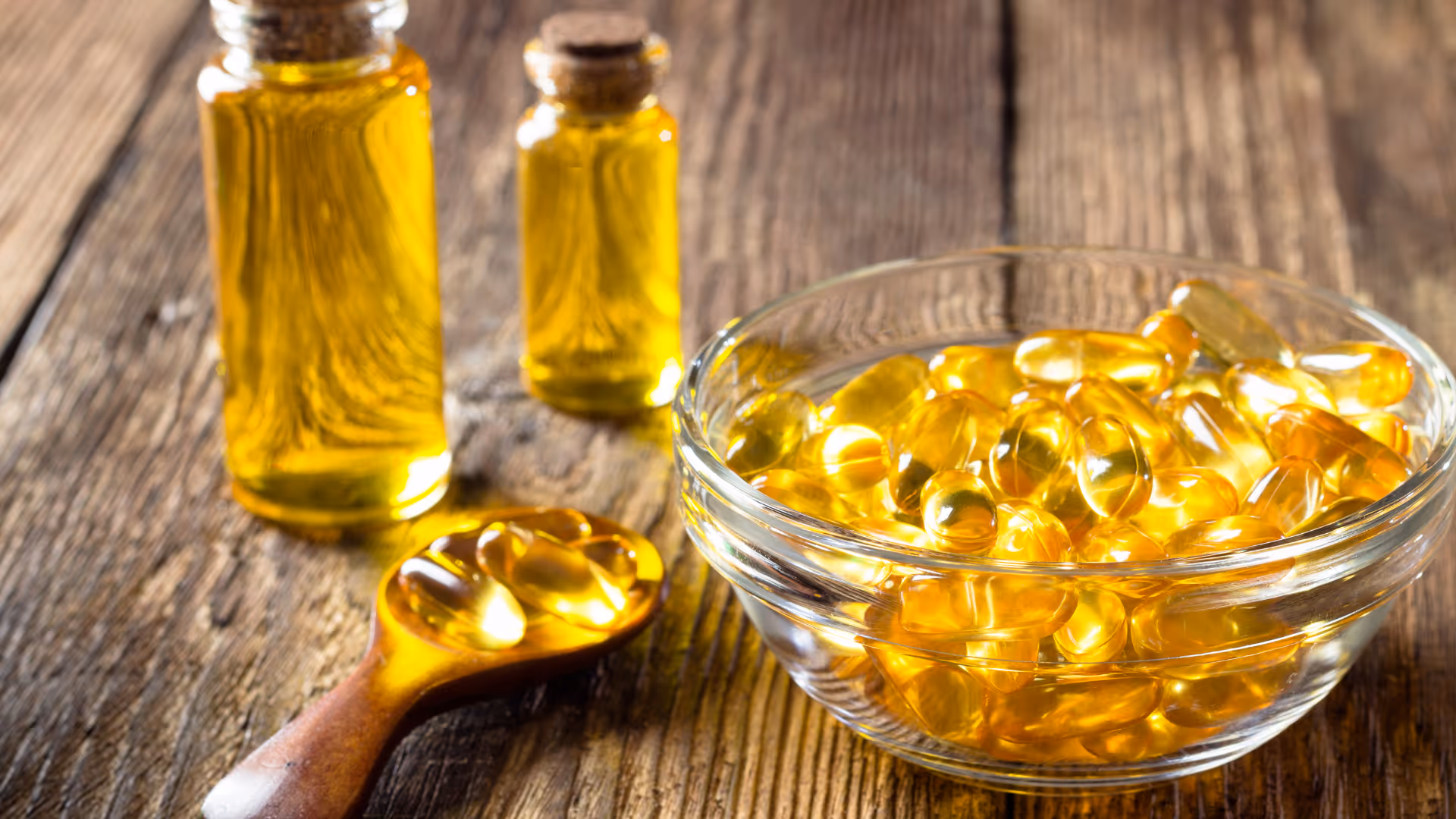 Schüssel und Löffel mit goldgelben Omega-3-Kapseln und zwei Flaschen mit Fischöl auf Holzuntergrund.