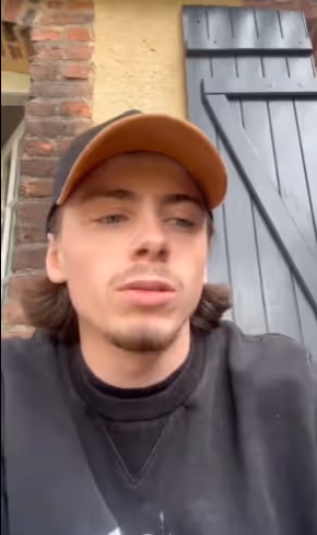 Young man with shoulder-length brown hair wearing a brown cap and black sweatshirt, sitting outdoors near a brick wall and a blue wooden door.