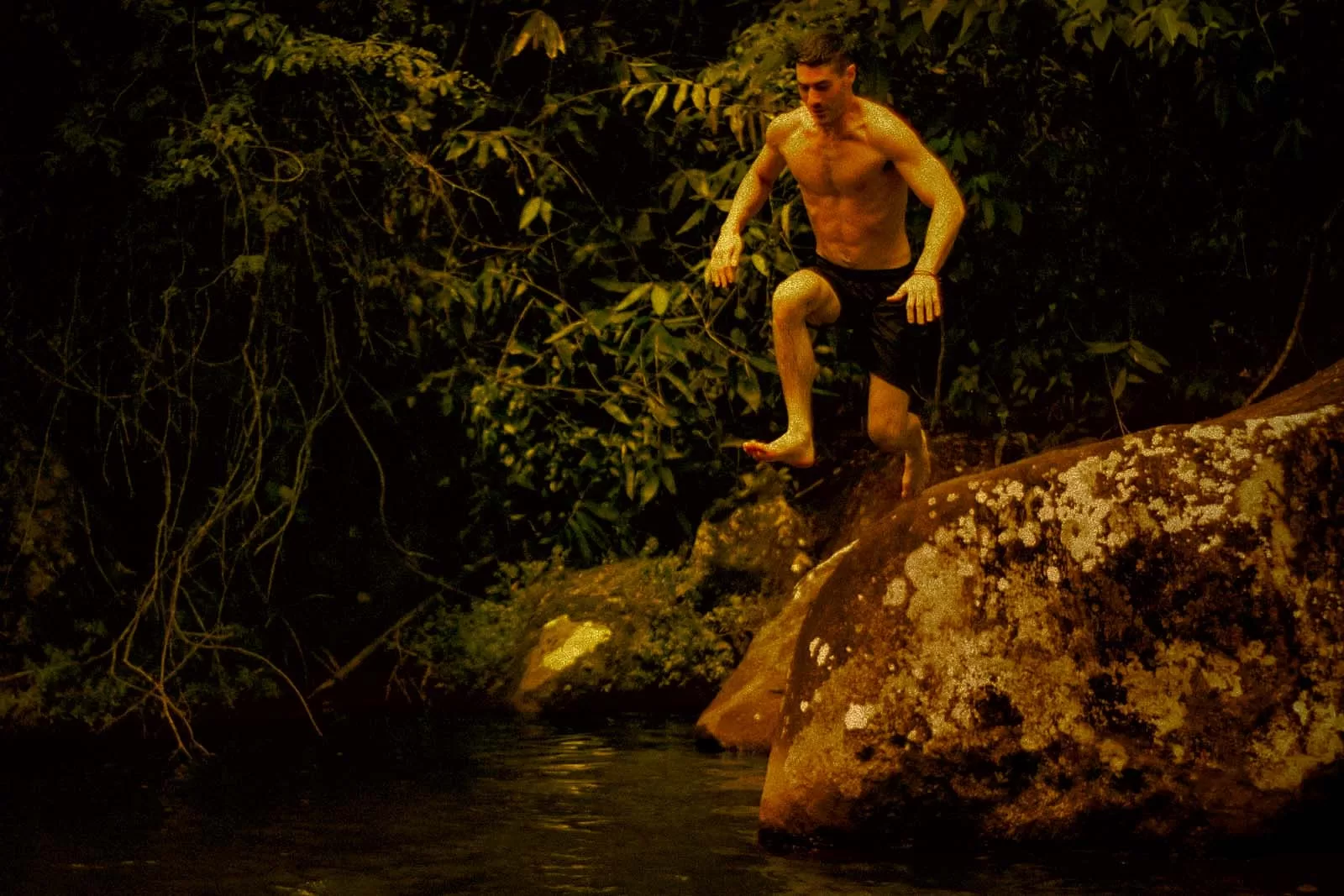 Shirtless man jumping off a large mossy rock into a dark river surrounded by dense foliage.