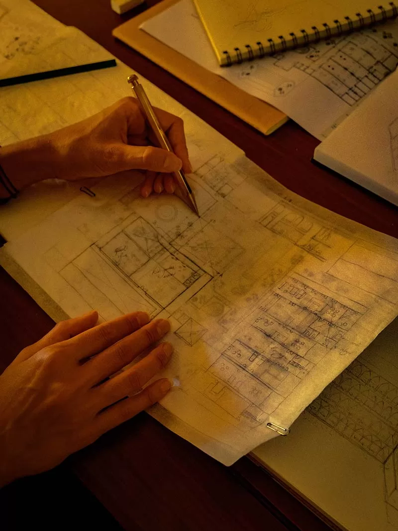 Person sketching architectural floor plans on tracing paper with notebooks and drawings on a wooden desk.