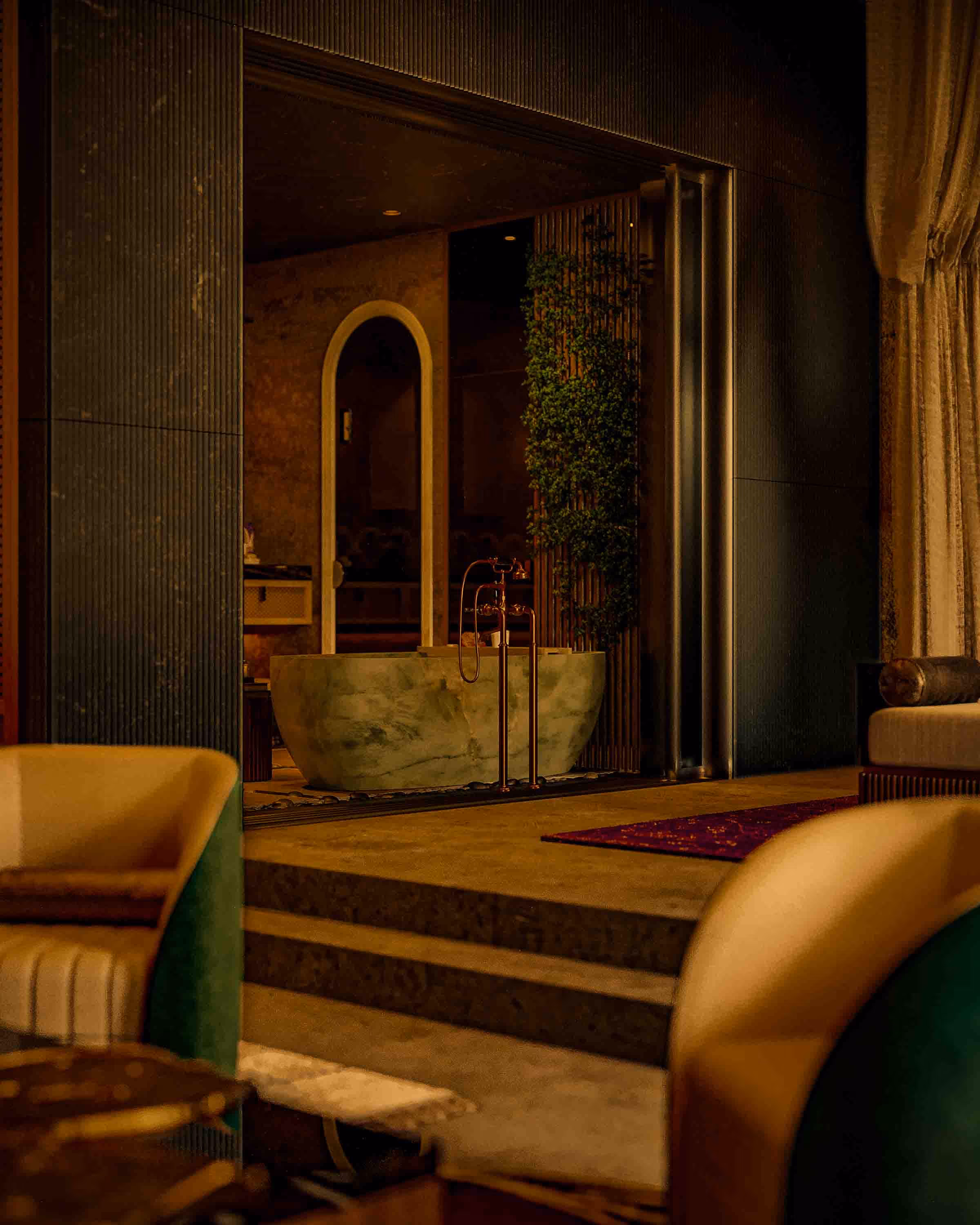 Luxurious bathroom with a stone bathtub, modern faucets, greenery on a wooden panel, and cozy armchairs in warm lighting.