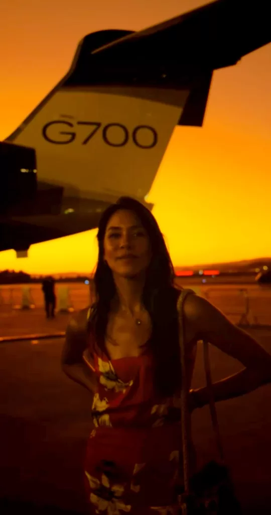 Woman in a red floral dress standing near a G700 private jet during sunset at an airport.