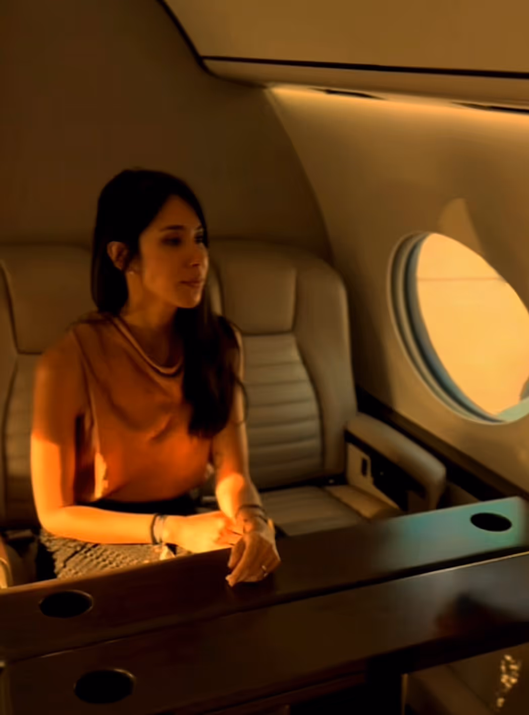 Woman seated at a wooden table inside a private jet with beige leather seats and round window.