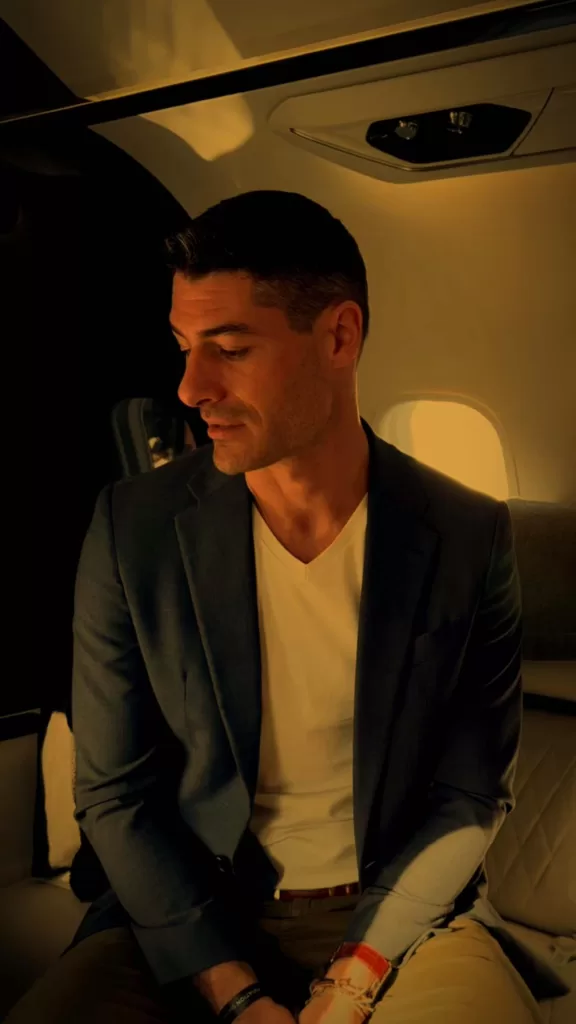 Man in a dark blazer and white shirt sitting inside a private jet with sunlight through the window.