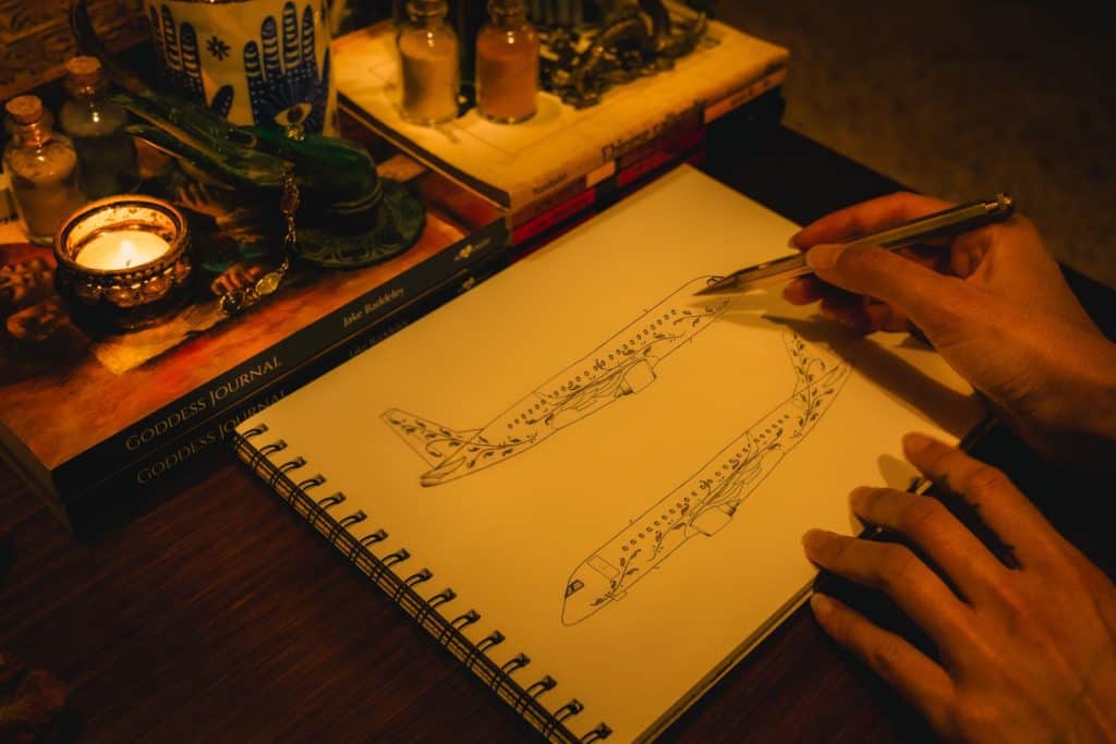 Person sketching detailed floral designs on two airplane illustrations in a spiral-bound notebook, next to lit candle and books on a wooden table.