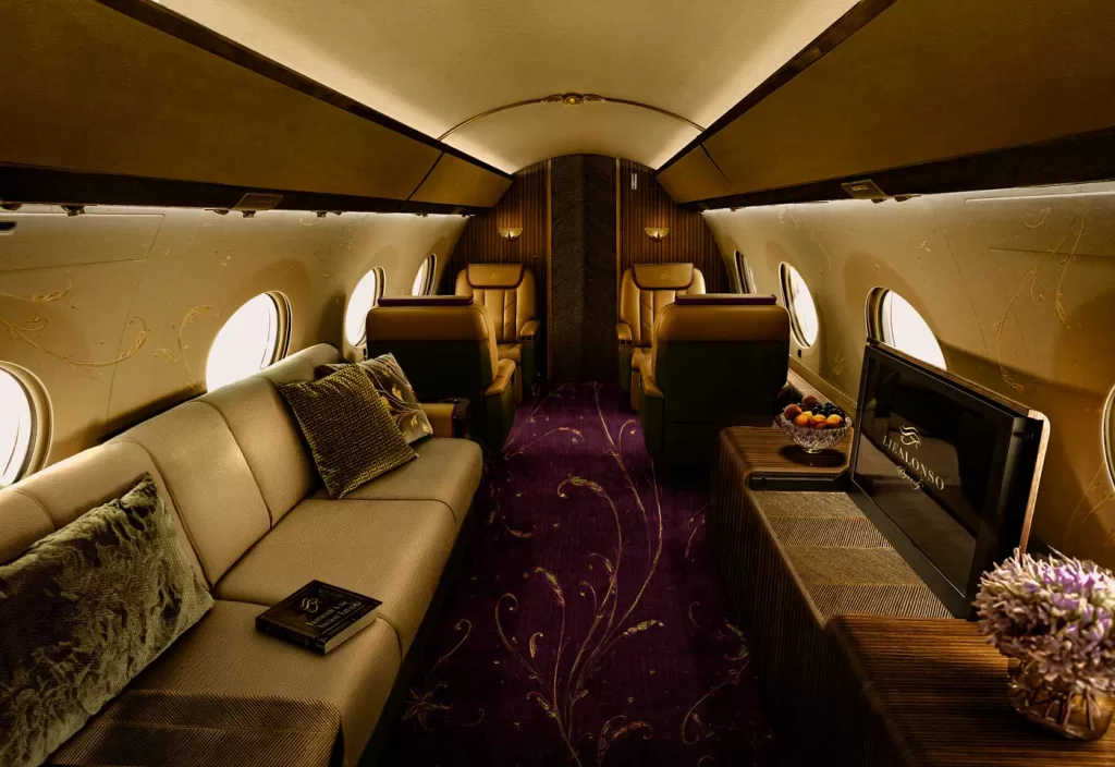 Luxurious private jet interior with beige sofa, leather seats, patterned purple carpet, and flat-screen TV.