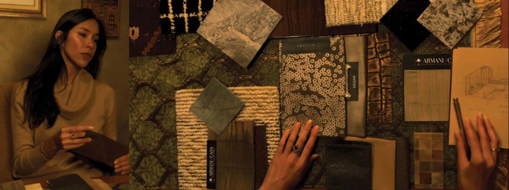 Woman seated at a table with various fabric and material samples spread out, holding one sample while reviewing design sketches.