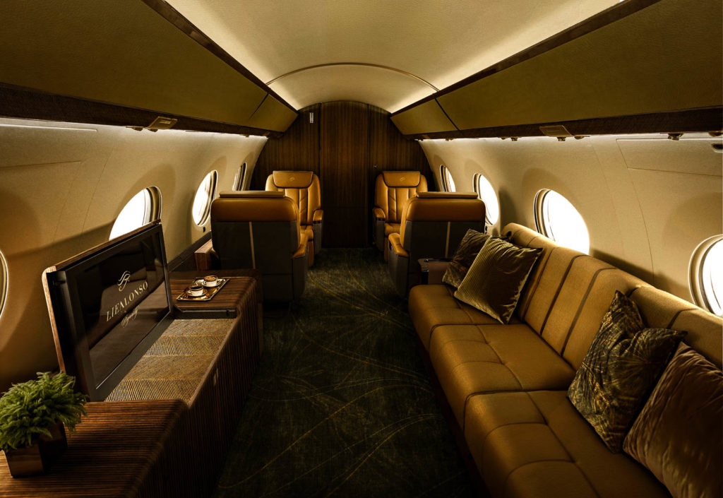 Luxurious private jet interior featuring tan leather seating, a long couch with pillows, a built-in TV, and wooden accents.