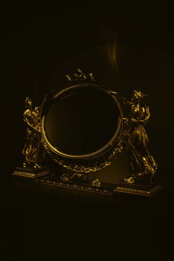 Ornate golden antique mirror held by two classical female statues on a dark background.