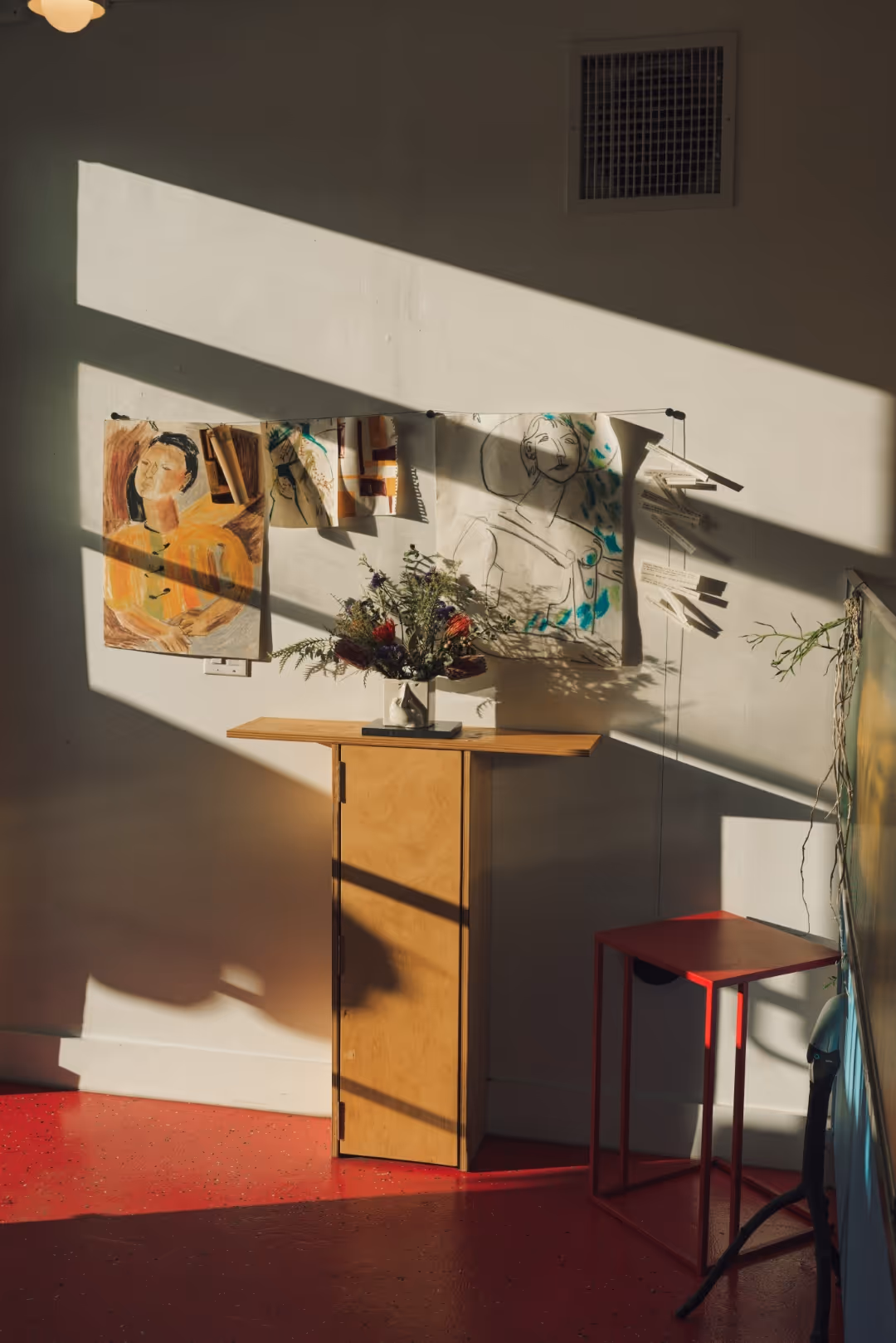 Sunlit corner with a small wooden cabinet holding a vase of flowers, a red metal stool, and sketches of faces pinned to the wall.