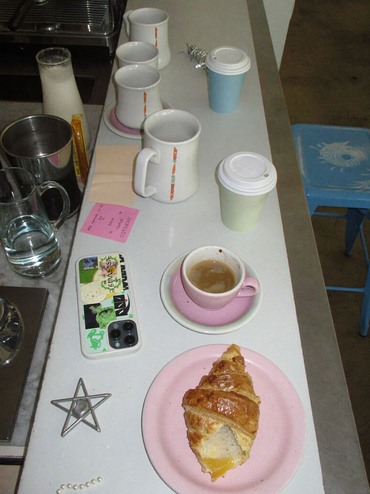 Mismatched mugs, espresso cups and a croissant lined up on a cafe counter