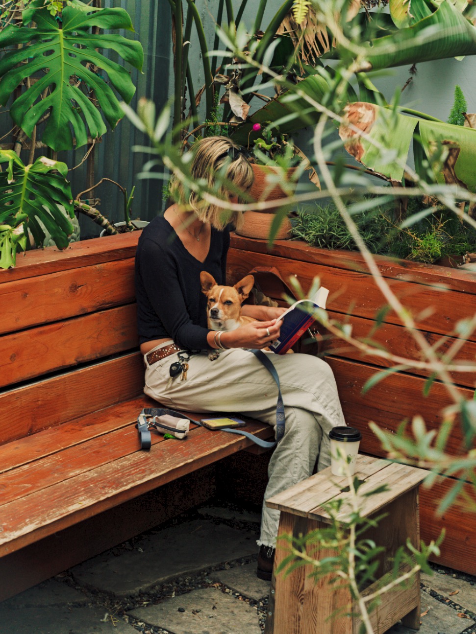 Woman reading with a small dog on her lap on a wooden bench surrounded by plants