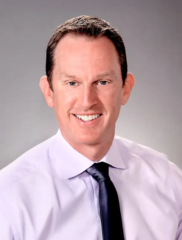 Professional headshot of smiling man in light purple shirt and dark tie.