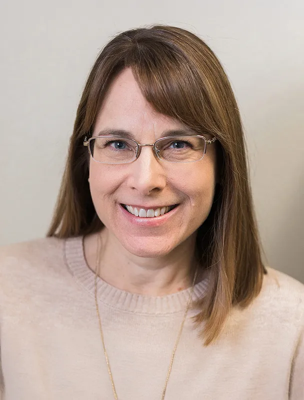 Professional headshot of smiling woman wearing glasses and beige sweater.