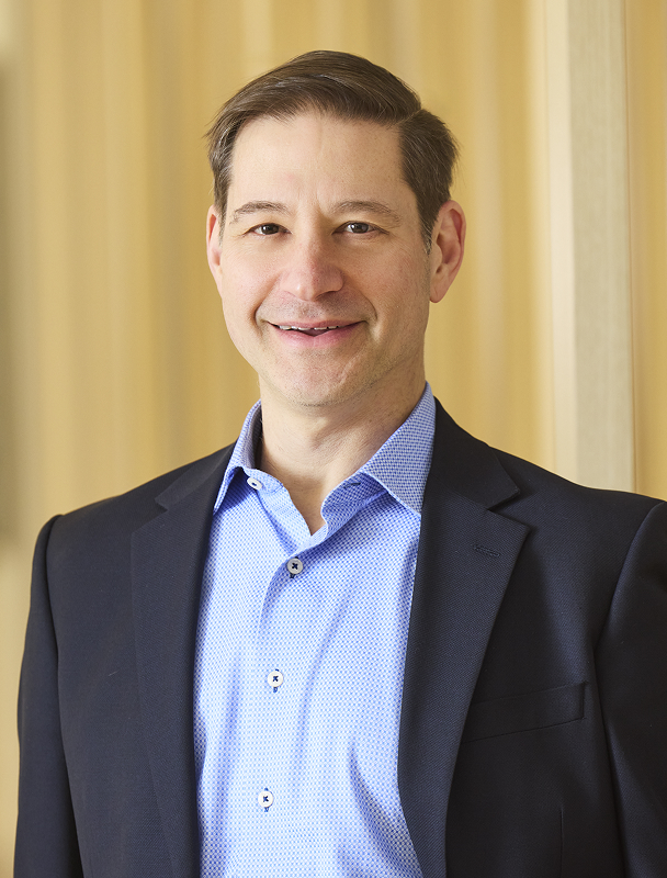 Professional headshot of smiling man in blazer and button-down shirt.