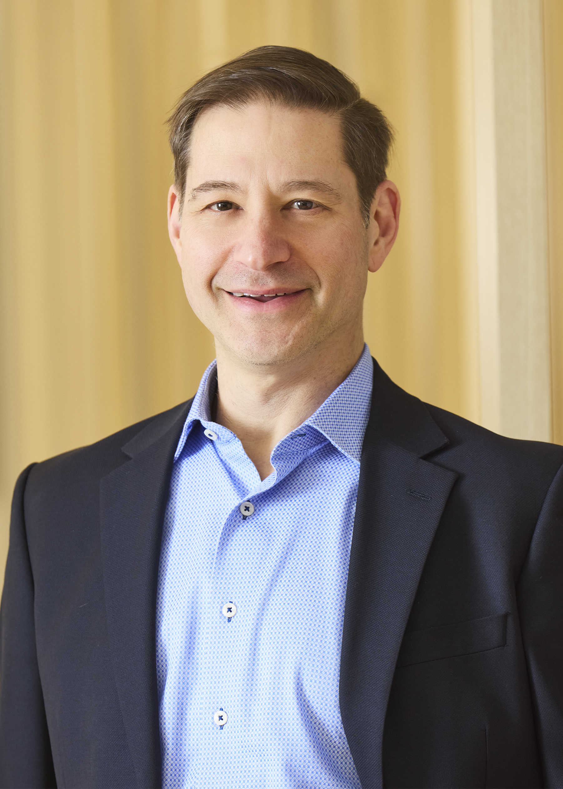 Professional headshot of smiling man in blazer and button-down shirt.