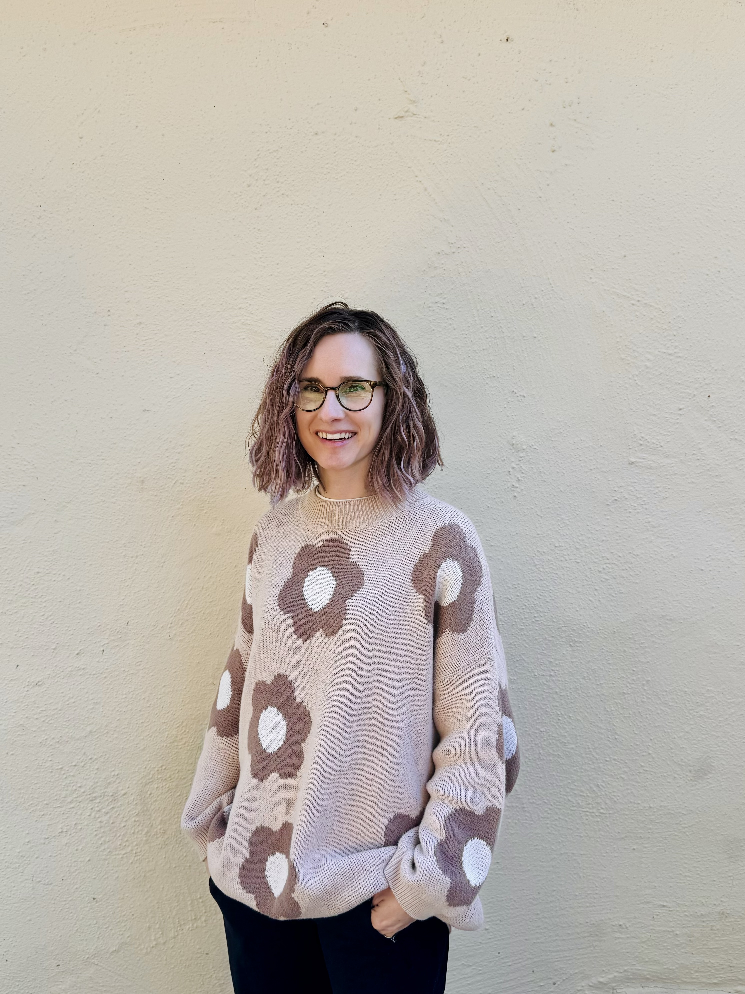An image of Amanda in a flower sweater  standing and smiling in front of a blank wall