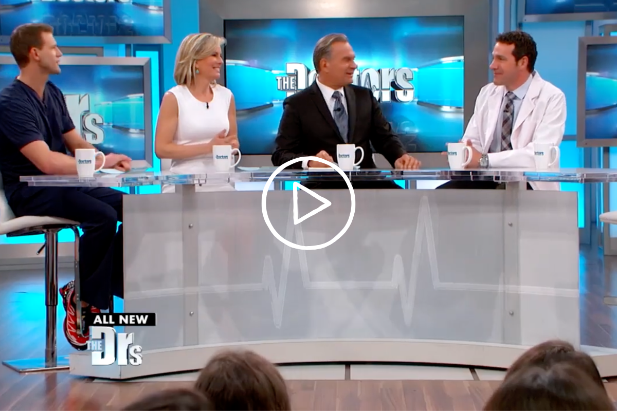 Four hosts seated behind a transparent desk on the set of The Doctors TV show, smiling and conversing.