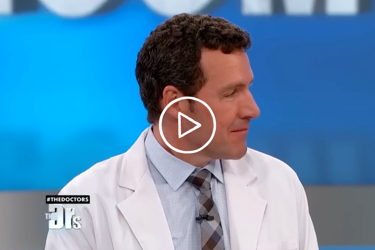 A man with curly dark hair wearing a white lab coat, light blue shirt, and patterned tie looking to the side on a medical TV show set.