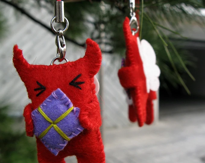 Felt ornaments in the shape of little red devils with angel wings. Each devil is clutching a purple wrapped present.