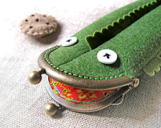 Felt pencil case in the shape of a green alligator, next to a brown felt chocolate chip cookie lapel pin..