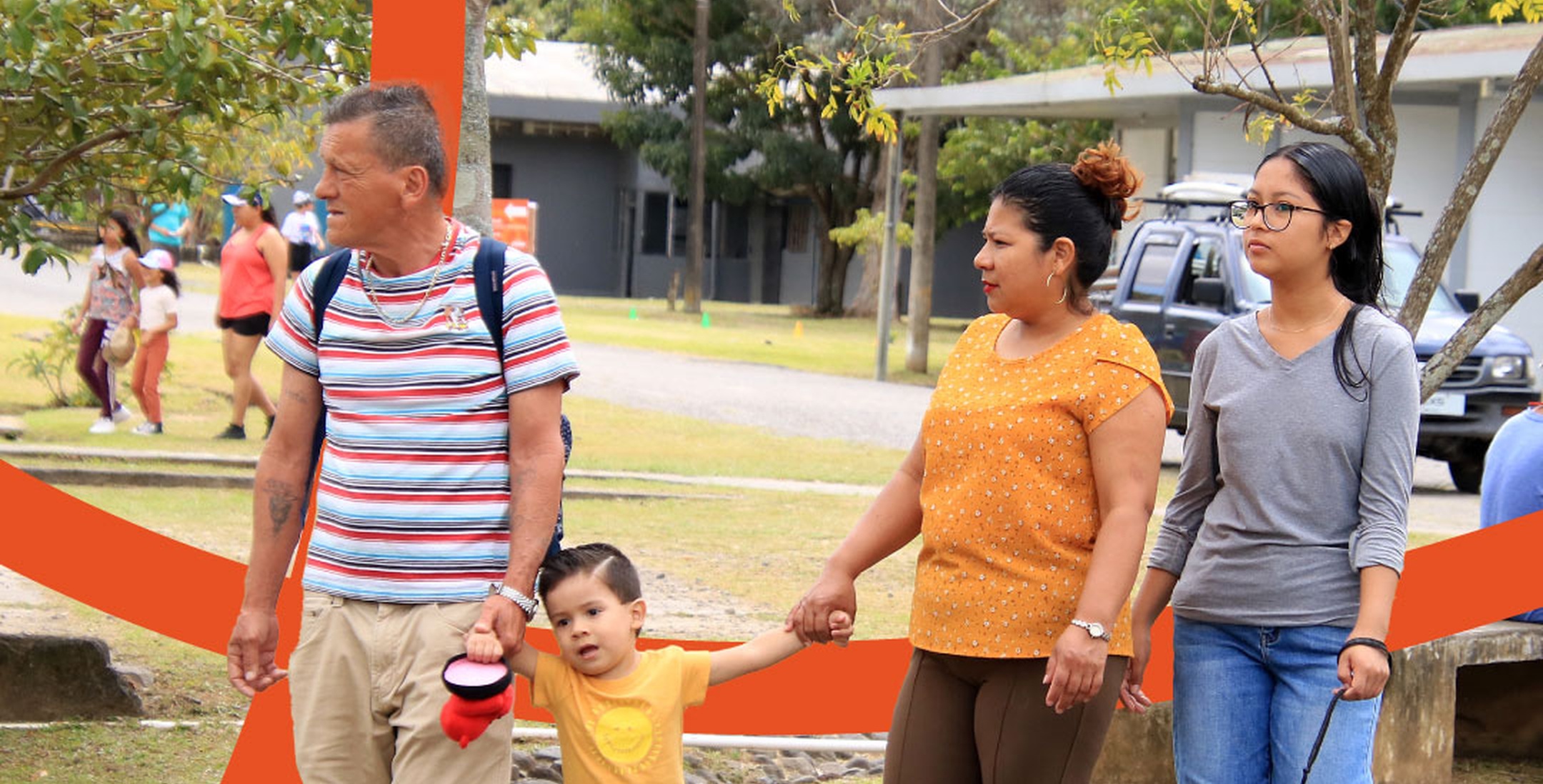 Family of four walking outside, with father, mother, young boy, and teenage girl holding hands in a park-like setting.