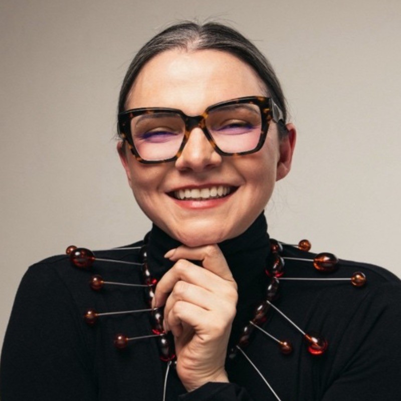 Smiling woman with glasses wearing a unique necklace with large beads and pins on a black turtleneck.