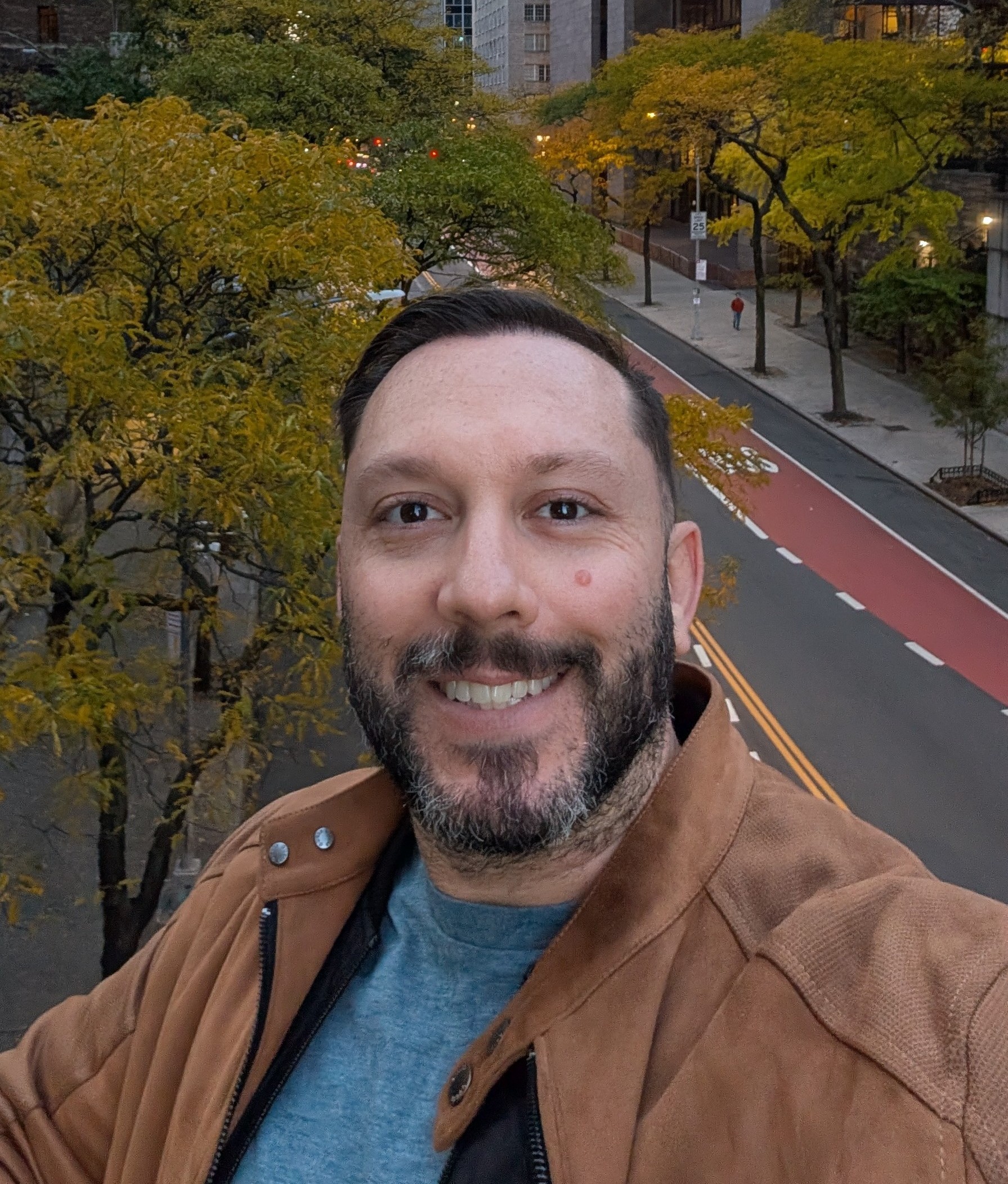 Smiling man with beard wearing brown jacket and blue shirt outdoors near a street with fall trees.