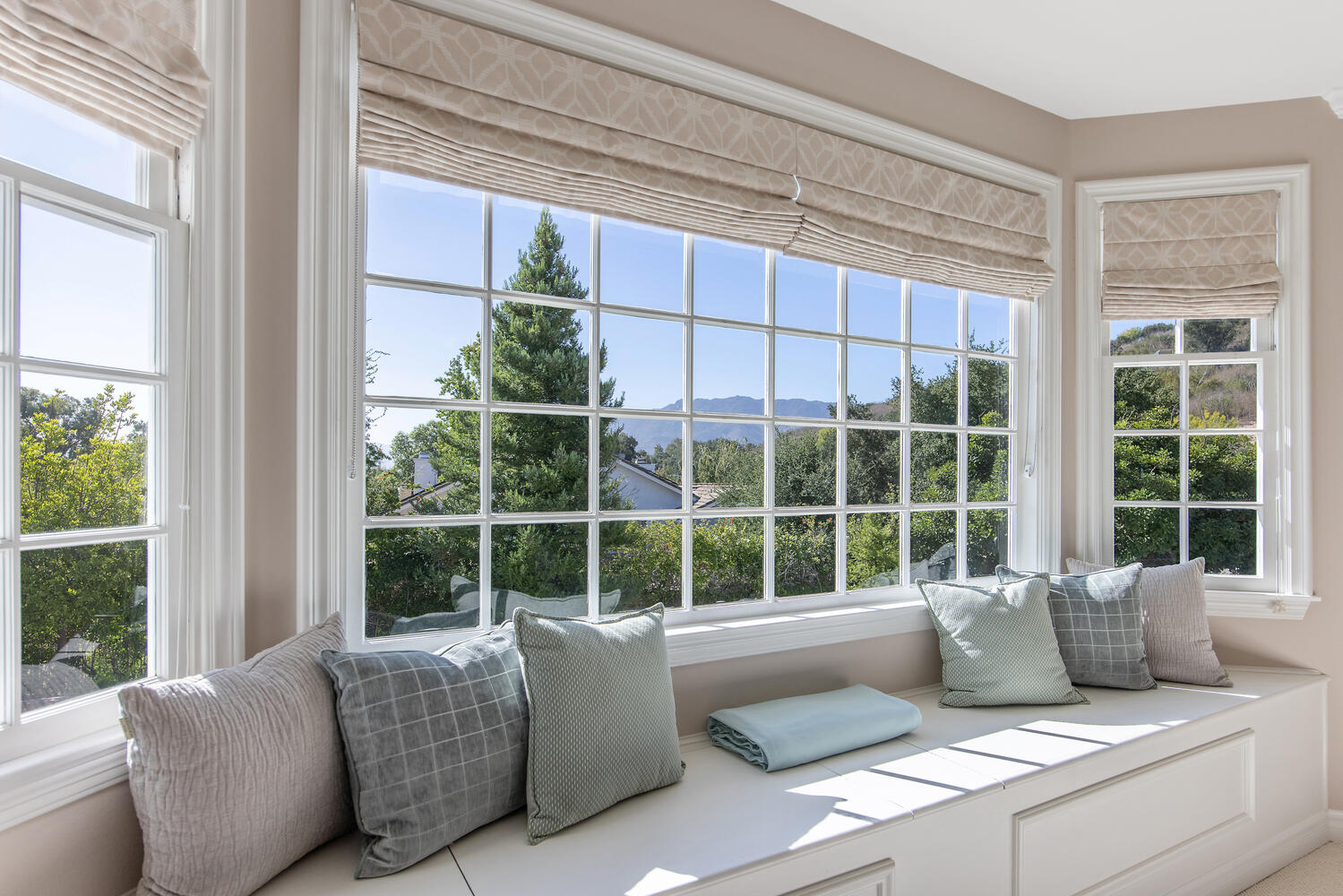 Sunlit window seat with six decorative pillows and a folded blanket, overlooking trees and mountains outside.