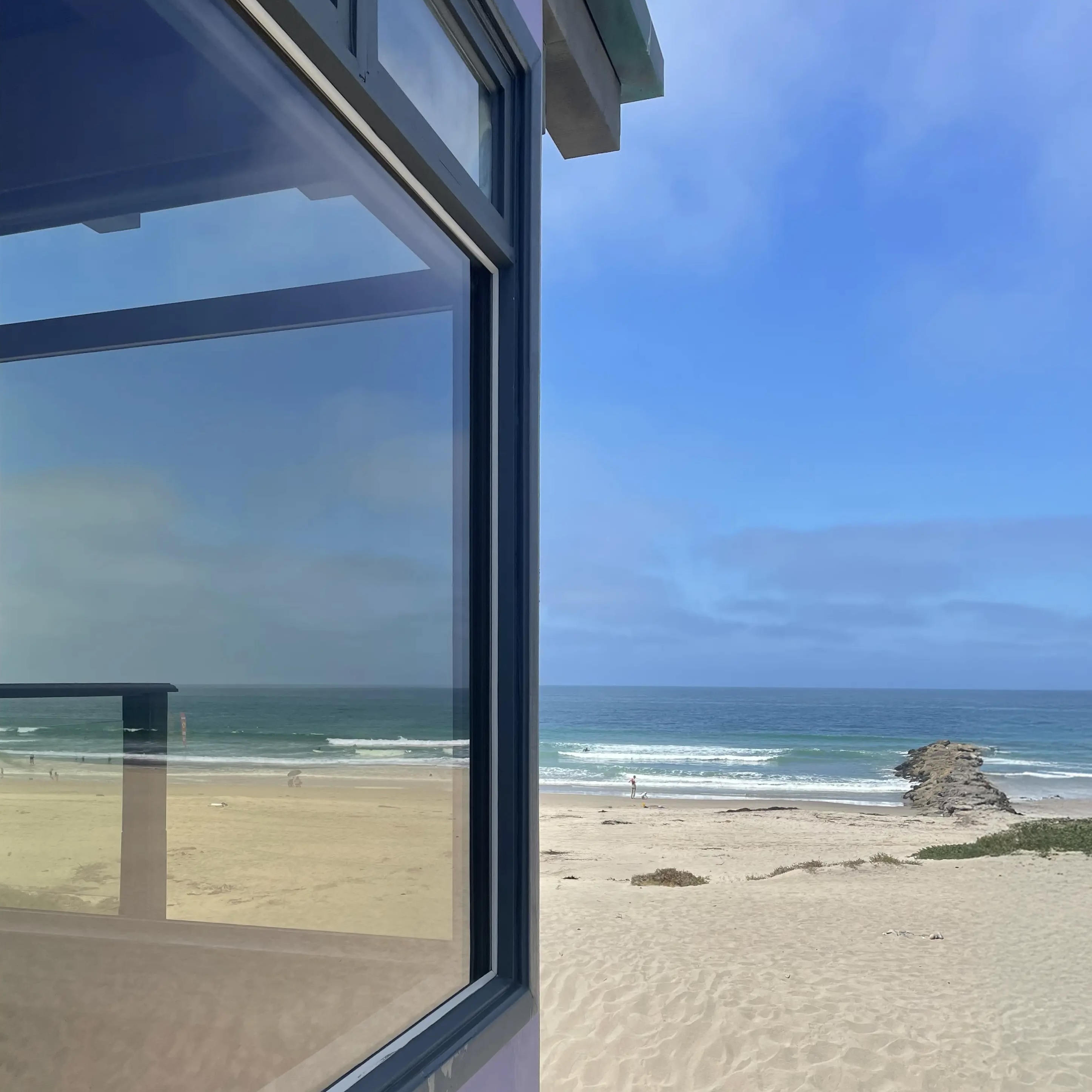 View of a sandy beach with gentle waves and a rocky jetty, seen next to a large window reflecting the sky.