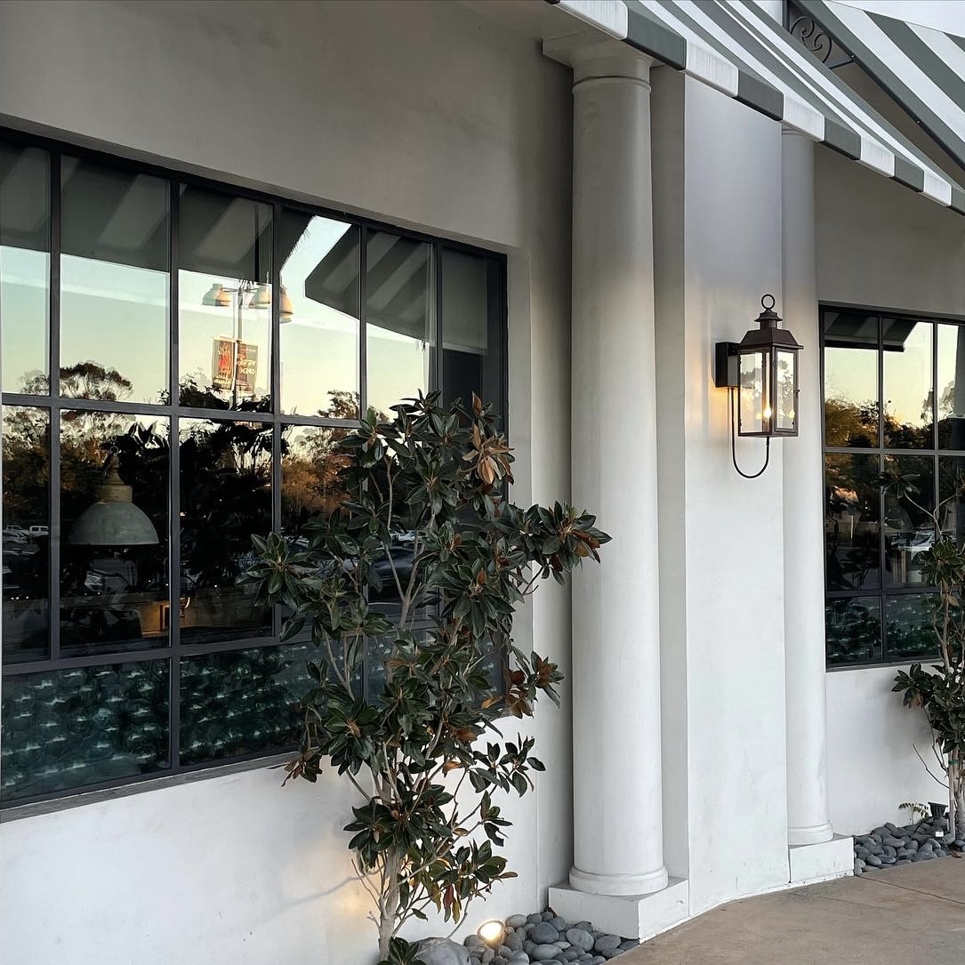 Exterior view of a building with white columns, black-framed windows, a wall-mounted lantern, and plants surrounded by river rocks.